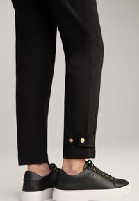 Zwarte tapered broeken met zichtbare naden en gouden knoopaccenten op de boorden, gecombineerd met zwarte leren sneakers met een witte zool.