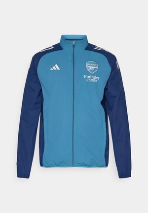Veste zippée bleu et marine avec le logo d'Arsenal, le marquage "Emirates" et des bandes blanches Adidas sur les épaules. Texture lisse, coupe athlétique.