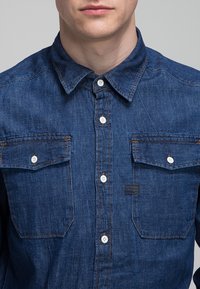Homme portant une chemise en jean bleu à boutons avec deux poches poitrine et des boutons blancs, visible du cou jusqu'au milieu du torse.