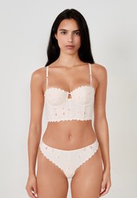 Completo di lingerie rosa pallido con bordi ondulati; presenta dettagli ricamati. Il top ha un design strutturato, mentre il fondo è un perizoma abbinato.