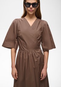 Femme portant une robe portefeuille marron à manches larges et des lunettes de soleil carrées noires, debout devant un fond blanc uni.