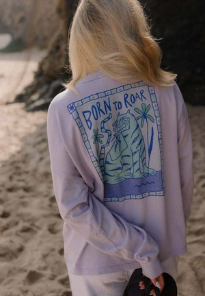 Blondynka w jasnofioletowej bluzie z kapturem z niebiesko-zielonym tygrysem i napisem "Born to Roar" na plecach, stojąca na piaszczystym podłożu.