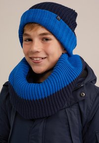 Blå och marinblå stickad mössa och matchande oversized halsduk, med ribbad textur och tjockt, mysigt tyg, bärs med en mörk kappa.