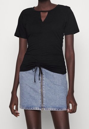 Femme portant un haut noir à manches courtes froncé avec un décolleté en trou de serrure et une mini-jupe en jean bleu clair avec une fermeture éclair à l'avant.