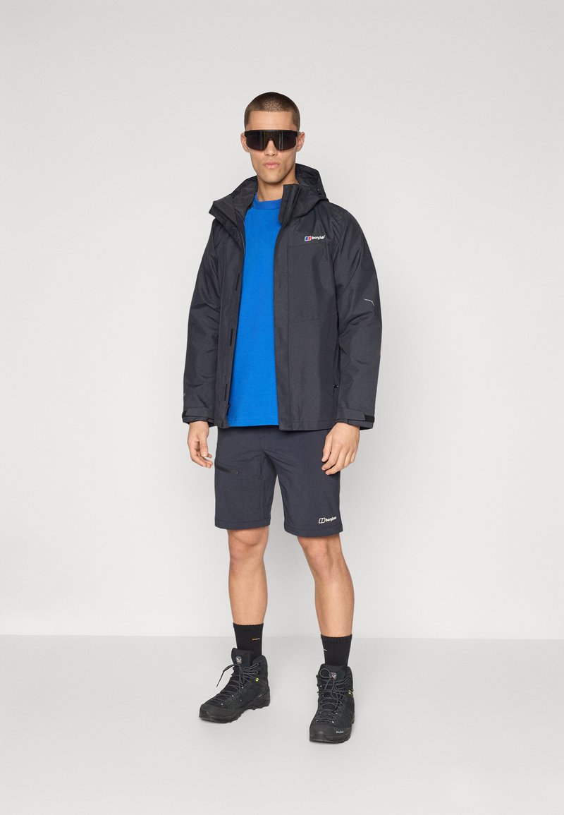 Czarna wodoodporna kurtka z kapturem, niebieska koszula, ciemne krótkie spodenki i czarne wodoodporne buty. Zawiera logo i odblaskowe akcenty.