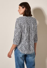 Camicia bianca con un motivo a foglia nera, maniche a tre quarti e orlo arrotondato, abbinata a jeans in denim blu.