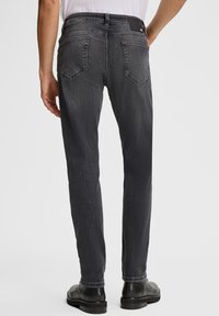 Uomo che indossa jeans slim-fit grigio scuro e scarpe di pelle nere, mostrato di spalle contro uno sfondo chiaro e uniforme.