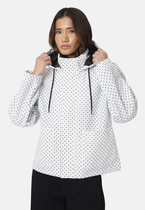 Femme ajustant la capuche d'une veste blanche à pois noirs avec des cordons noirs, portant un pantalon noir sur un fond uni.