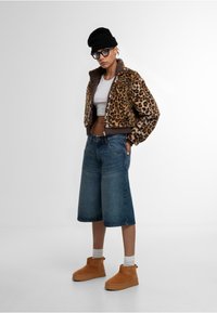 Leopárdmintás műszőrme dzseki, fehér crop top, kék farmer culotte nadrág, barna textúrás felületű bokacsizma, fekete sapka és szemüveg.