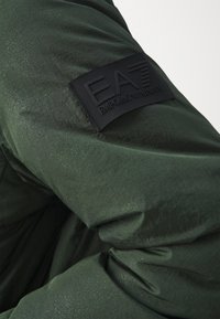 Zelena pufasta jakna s dugim rukavima izrađena od glatkog, sjajnog materijala, s teksturiranim crnim ušivenim dijelom i EA7 Emporio Armani logotipom.