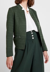 Femme portant un blazer texturé vert foncé, une chemise blanche à boutons, et un pantalon taille haute vert foncé avec quatre boutons visibles.