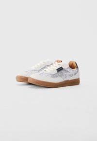 Zapatillas bajas blancas y plateadas con suela de goma, cordones beige y una lengüeta negra en el talón, colocadas una al lado de la otra sobre un fondo blanco.