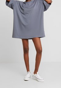 Person i en oversize grå T-shirt og hvide lavtoppede sneakers, stående mod en ensfarvet lys grå baggrund.