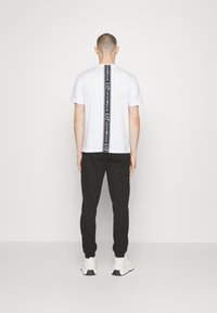 Camiseta blanca de manga corta con un detalle de logo vertical en blanco y negro en el centro, combinada con pantalones negros y zapatillas blancas.
