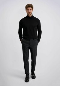 Man in een zwarte coltrui, donkergrijze pantalon en zwarte dresschoenen, staand met handen in zijn zakken tegen een effen achtergrond.