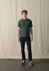 T-shirt verte à manches courtes avec un col rond, associée à un pantalon sombre et des baskets multicolores. Sur fond neutre.