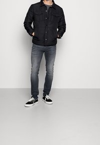 Svart puffjacka med krage och tryckknappar, parat med slim-fit ljusgrå jeans och svarta sneakers med vita sålar.