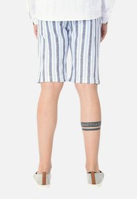 Bermuda in lino a righe blu e bianche, con un tatuaggio sulla gamba inferiore. La persona indossa scarpe slip-on grigie con un accento marrone.