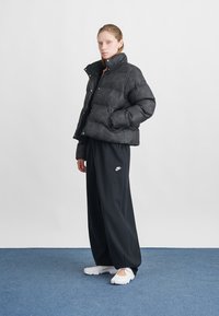 Čierna prešívaná bunda s vysokým golierom, prešívaným dizajnom a cvokmi, skombinovaná so voľnými čiernymi nohavicami s logom Nike a bielymi topánkami.