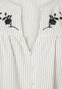 Witte stoffen blouse met zwarte verticale strepen en bloemenborduursel op de buste. Bevat knopen en geplooide details onder de borst.