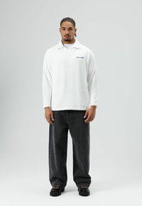 Witte, gestructureerde fleece trui met een kraag en een blauw geborduurd logo, gecombineerd met loszittende zwarte spijkerbroek en zwarte schoenen.