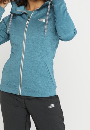 Person iført teal lynlås-hættetrøje og sorte bukser med The North Face-logoer, der justerer hættetrøje-kraven.