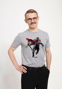 Camiseta de manga corta gris con un gráfico de un héroe con una capa roja y un hacha, impresa en negro y rojo. Se llevan pantalones negros y un cinturón.