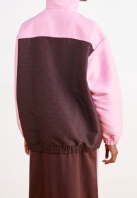 Färgbreakjacka med en rosa övre del och en brun nedre del med ett geometriskt mönster. Elastisk midja och ärmslutar.