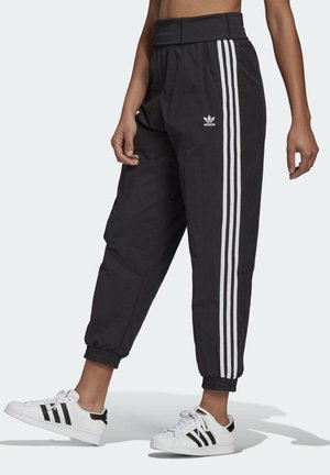 Osoba ubrana w czarne spodnie Adidas z białymi paskami i logo, w połączeniu z białymi butami sportowymi Adidas z czarnymi paskami, stojąca na jasnym tle.