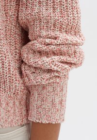 Neulepusero vaaleanpunaisessa ja kermassa, jossa on käännetyt hihansuut ja tekstuuri kuvioita. Materiaaleihin kuuluu lankaa, jossa on vivahteita kimalteesta.