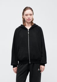 Svart hettegenser med glidelås, frontlommer og ribbede mansjetter og nederkant. Laget av teksturert stoff med en subtil logo på brystet.