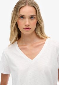 Mujer rubia con ojos azules, vistiendo una camiseta blanca de cuello en V, mirando directamente al frente contra un fondo blanco liso.