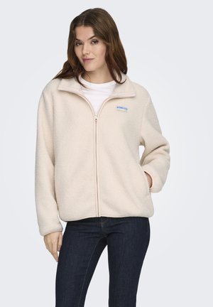 Woman wearing cream-colored fleece jacket with zipper, white shirt, and dark jeans, standing with one hand in pocket against white background.