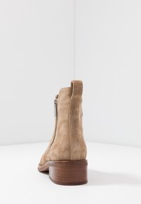 Beżowy zamszowy botek za kostkę o gładkiej fakturze, z tylnym zamkiem błyskawicznym i brązowym, warstwowym obcasem. Minimalistyczny design, bez widocznych wzorów czy zdobień.