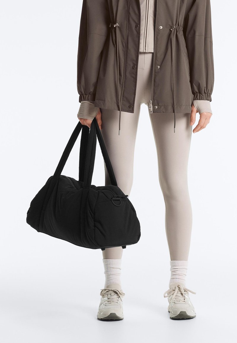 Person wearing taupe leggings, taupe jacket, white socks, and sneakers, holding a large black duffel bag by the handles.