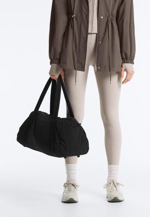 Person iført taupe leggings, taupe jakke, hvide sokker og sneakers, der holder en stor sort duffelbag i håndtagene.