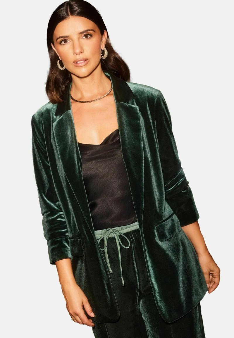 Blazer in velluto verde scuro con una vestibilità rilassata, rever a punte, tasche frontali, abbinato a un top in satin nero e pantaloni coordinati.