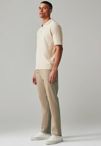 Kurzärmliges cremefarbenes Polohemd mit strukturiertem Muster, kombiniert mit hellbeigen Hosen und weißen Sneakers. Stehendes Modell im Profil.
