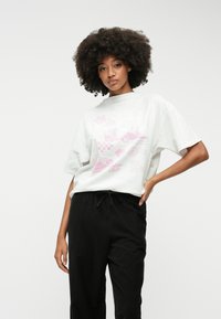 Fehér, rövid ujjú póló, amelyen rózsaszín grafikai mintázat látható, különféle mintákkal. Fekete, zsinóros derekú, bő szárú nadrággal párosítva.