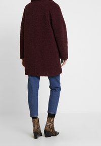Cappotto borgogna con texture e vestibilità rilassata, abbinato a jeans in denim blu e stivali alla caviglia in stampa pitone con tacco squadrato.