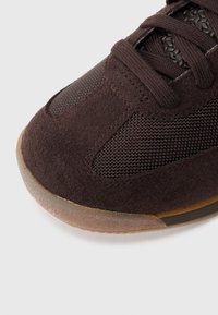 Hnědý teniska z broušené kůže a síťoviny s kulatou špičkou, texturovanou tkaninou, výrazným šněrováním a podrážkou z kaučukové gumy. Detailní pohled zdůrazňující detaily.