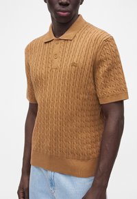 Homme portant un polo en maille torsadée couleur camel à manches courtes avec deux boutons et un jean bleu clair sur un fond uni.