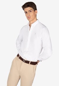 Camisa blanca de botones con mangas largas y un pequeño logo bordado en el pecho izquierdo, combinada con pantalones beige y un cinturón marrón.