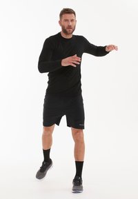 Schwarzes langärmliges Sportshirt kombiniert mit schwarzen Shorts. Das Modell trägt schwarze Socken und graue Sneakers mit blauen Akzenten in einer dynamischen Pose.