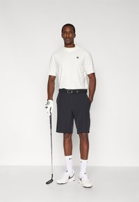 Biała koszula z krótkim rękawem o fakturze, ciemne szorty, czarny pasek, rękawica golfowa i kij golfowy. Noszone z białymi skarpetkami i butami. Neutralne tło.