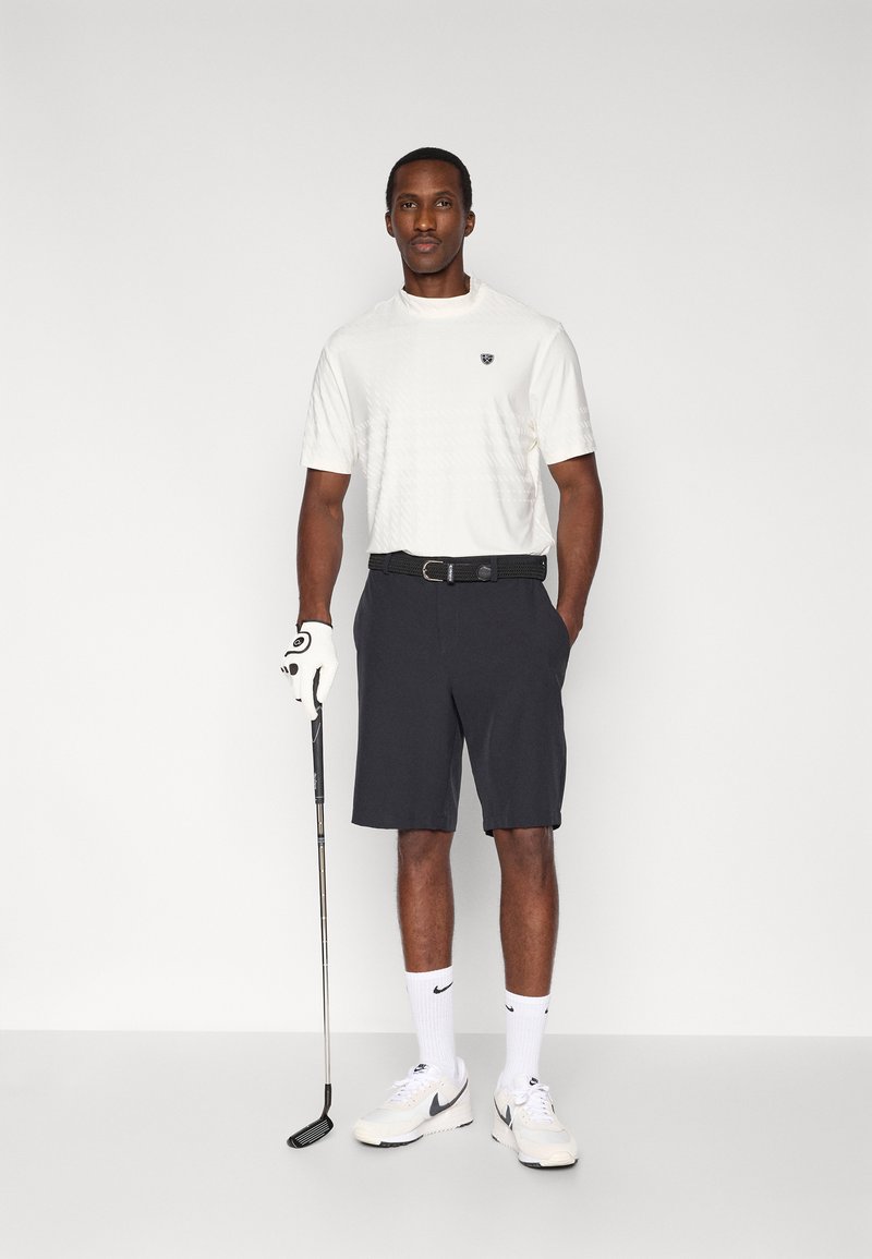 Camicia bianca a maniche corte con texture, pantaloncini scuri, cintura nera, guanto da golf e mazza da golf. Indossata con calzini e scarpe bianchi. Sfondo neutro.