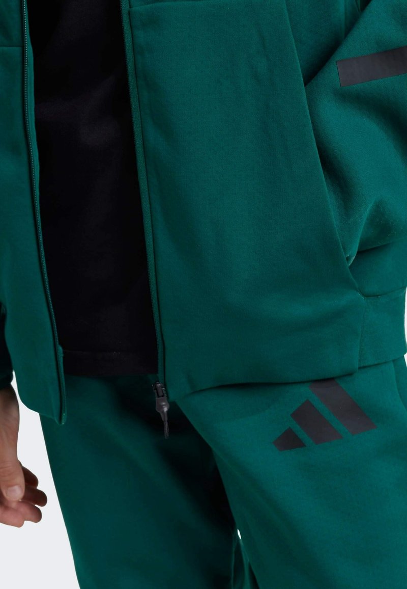 Grön sportoutfit med en jacka med dragkedja och byxor. Materialet har en mjuk textur med svarta detaljer och en tre-randig logotyp på byxorna.