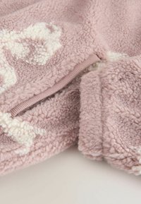Pehmeä vaaleanpunainen fleece-kangas, jossa on teksturoitu, pörröinen pinta. Sisältää valkoisen abstraktin kuvion ja ribattuja saumoja. Lähikuva.