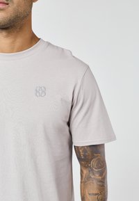 Bărbat cu cămașă cu mânecă scurtă, bej deschis, și tatuaj detaliat cu trandafir și balanțe pe antebraț, pe fundal simplu.