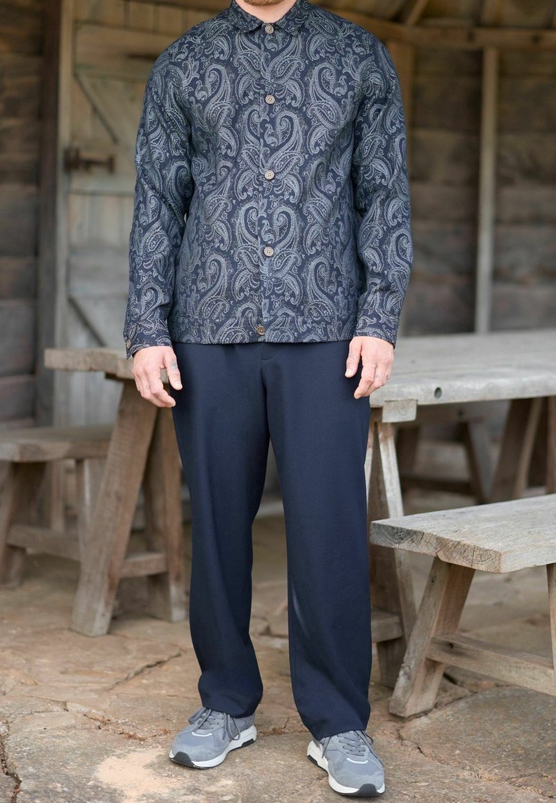 Homme portant une chemise à boutons à motif paisley foncé, un pantalon bleu marine et des baskets grises, debout à l'intérieur d'une structure en bois rustique avec des bancs.
